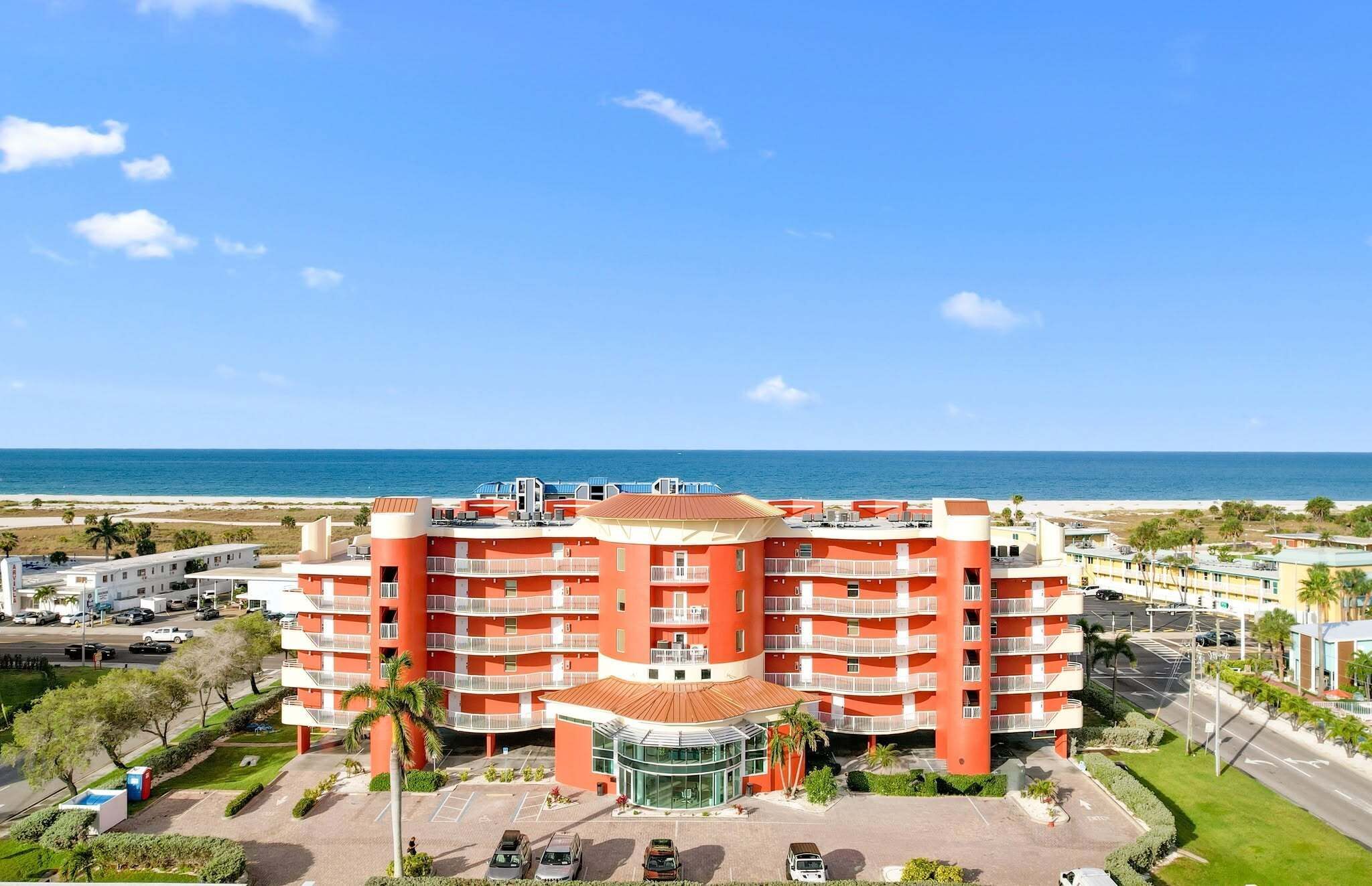A multi-story hotel with a central courtyard, surrounded by a parking area and a view of the beach.