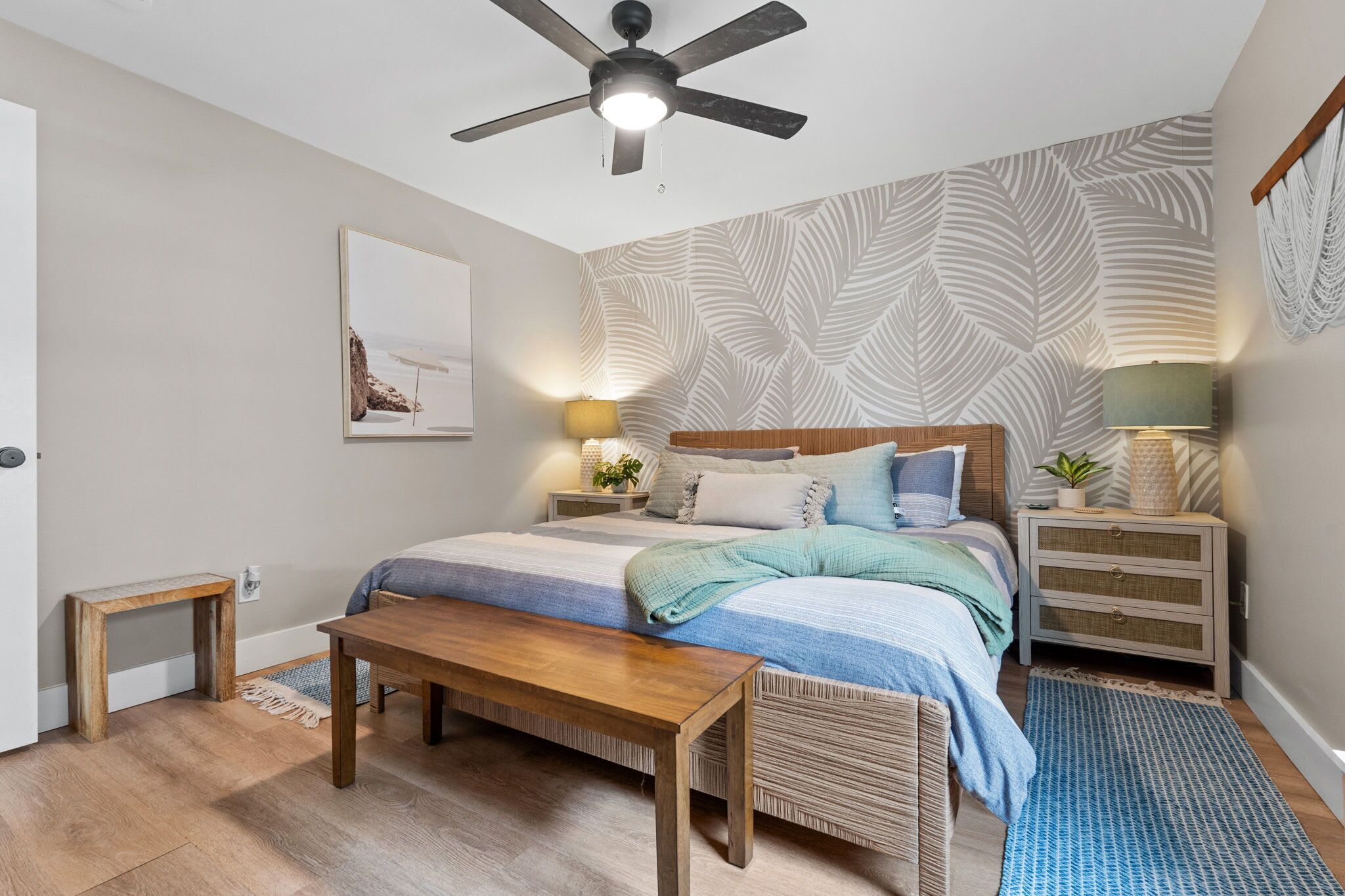 Un dormitorio con cama, mesitas de noche, ventilador de techo, un banco de madera, alfombra y un muro con estampado de hojas.
