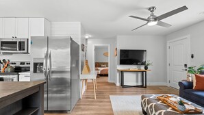 Una cocina moderna con electrodomésticos de acero inoxidable, un comedor con mesa y sillas, una sala de estar con sofá y televisor, y un ventilador de techo.