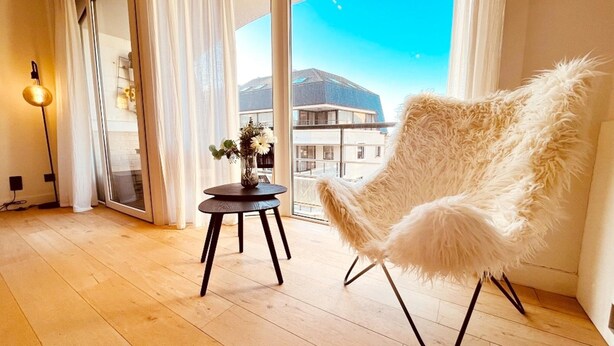 A room with a large window, a fluffy chair, a small table with flowers, and a lamp. - Apartment in Wenduine Near Zeedijk Beach (De Haan)