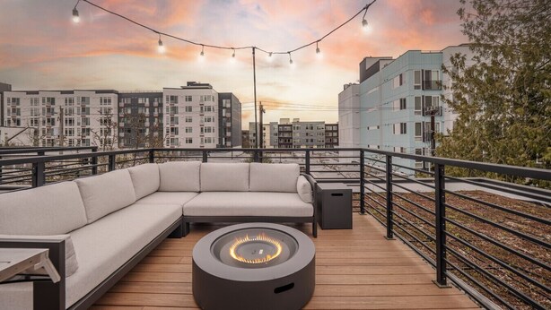 Terraza en la azotea con un área de estar exterior moderna, que incluye un sofá y un pozo de fuego circular, con vistas a un paisaje urbano al atardecer.