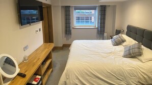 A bedroom with a bed, a TV mounted on the wall, a desk with a mirror, and a window with curtains. - Maypole Farm Guest House (Selby)