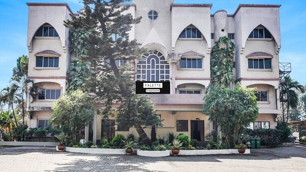 A multi-story building with a central arched entrance, surrounded by potted plants and trees. - Elegant Escape in Palette Bhimpore Daman (Daman)