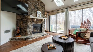 A living room with a stone fireplace, a flat-screen TV, a ceiling fan, and a large window with a view of trees. - ❄ The Snowballer – Ultimate Ski-In/Ski-Out Retreat! 🎿🏔 (Champion)