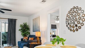 Ein modernes Wohnzimmer mit Essbereich, mit einem runden Tisch und Platzsets, einem blauen Sofa mit farbigem Kissen, einer großen Pflanze und einem Wandbild.