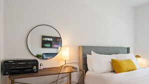 Ein Hotelzimmer mit einem Bett, einem Schreibtisch mit Drucker, einem runden Spiegel und einem an der Wand befestigten Fernseher, der Hotelkanäle zeigt.