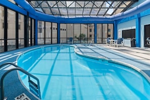 An indoor swimming pool with clear glass walls, a blue pool deck, and a covered roof structure.