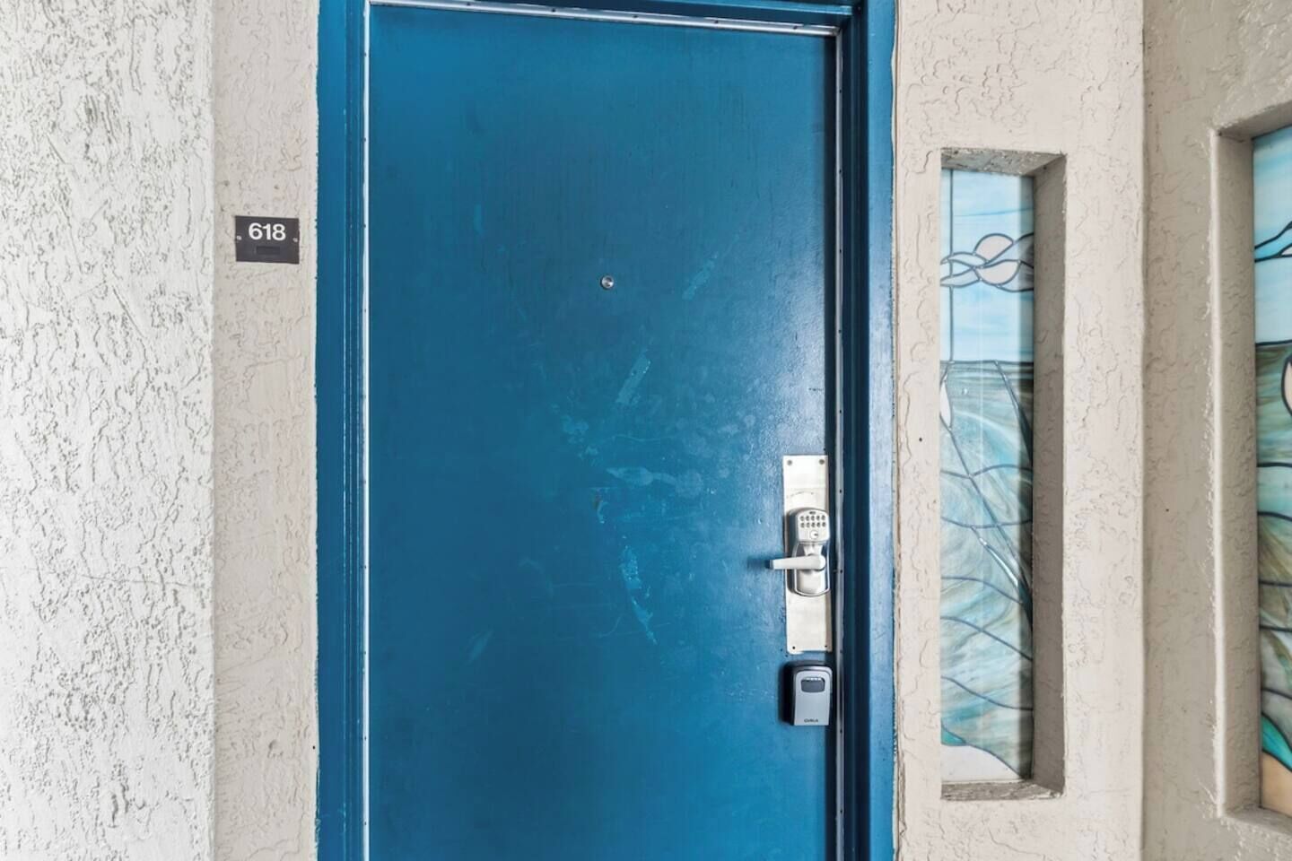 Une porte bleue dotée d’un clavier numérique, une petite fenêtre ornée d’un cadre décoratif et une plaque fixée au mur portant le numéro 618.