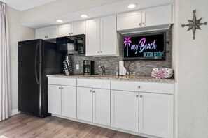 A modern kitchen with white cabinets, a black refrigerator, a microwave, and a coffee maker. There's a neon sign on the wall advertising vacation rentals. - Beachfront Haven | Ocean Views & Pool | Sleeps 4 (North Myrtle Beach)