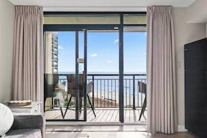 A balcony with a view of the beach and ocean, a chair, and a table with a lamp.