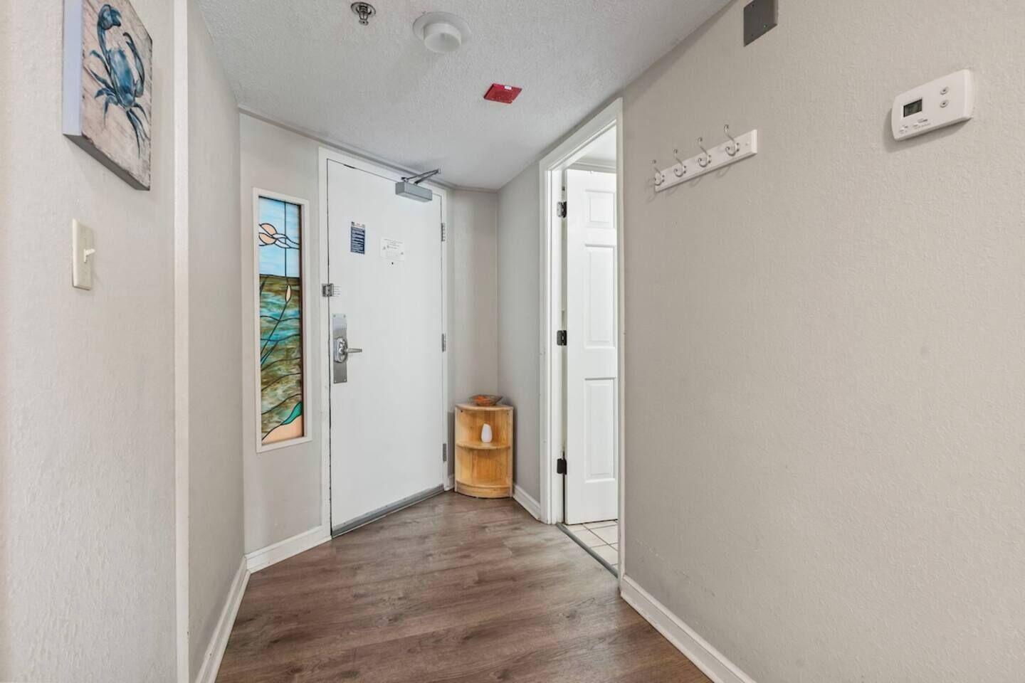 Un pasillo con suelo de madera, paredes blancas, una puerta con un vitral, un pequeño estante de madera y un termostato en la pared.