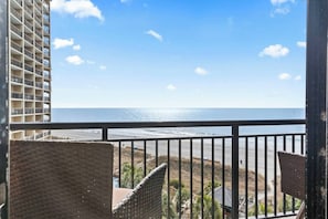 Ein Balkon mit Blick auf Strand und Meer, ein Gebäude im Hintergrund und ein Geländer.