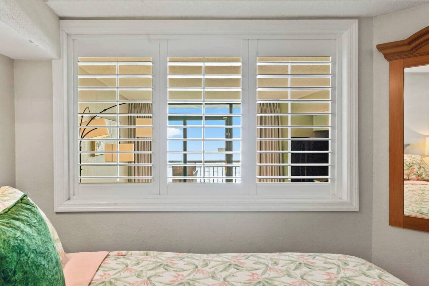 Une pièce avec une grande fenêtre, un lit recouvert d’une literie à motifs floraux et un miroir au mur.