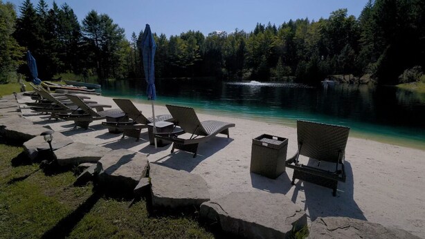Ein See-Loungebereich mit Liegestühlen und Sonnenschirmen, umgeben von Bäumen und mit freiem Blick auf den See.
