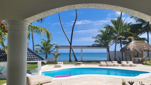 Un área de piscina con vista al océano, palmeras y una estructura de techo de paja.