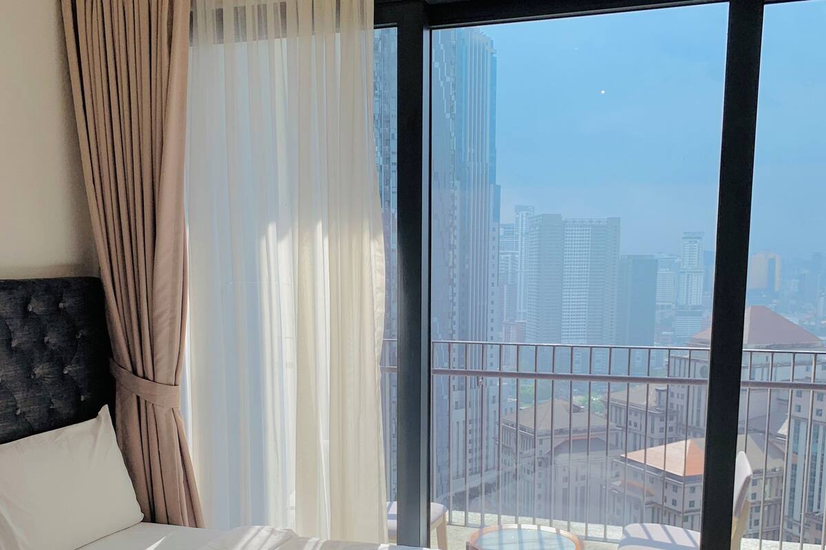 A hotel room with a large bed, a view of the cityscape through the window, and a balcony with a railing.