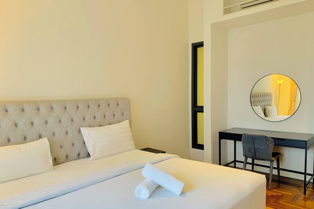 A neatly made bed with a tufted headboard, a round mirror on the wall, and a desk with a chair.