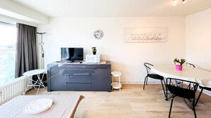 A modern living room with a flat-screen TV, a microwave, a dining table with chairs, a small side table, and a potted plant. - Studio in Wenduine Near the Beach (De Haan)
