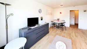 A modern living room with a flat-screen TV, a dining table with chairs, a white sofa, and a floor lamp. - Studio in Wenduine Near the Beach (De Haan)