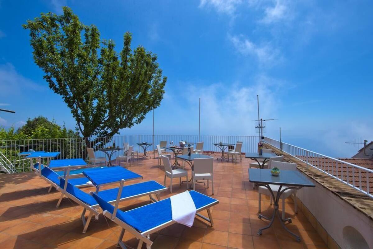 A rooftop terrace with blue lounge chairs, white tables, and chairs, surrounded by a metal railing.
