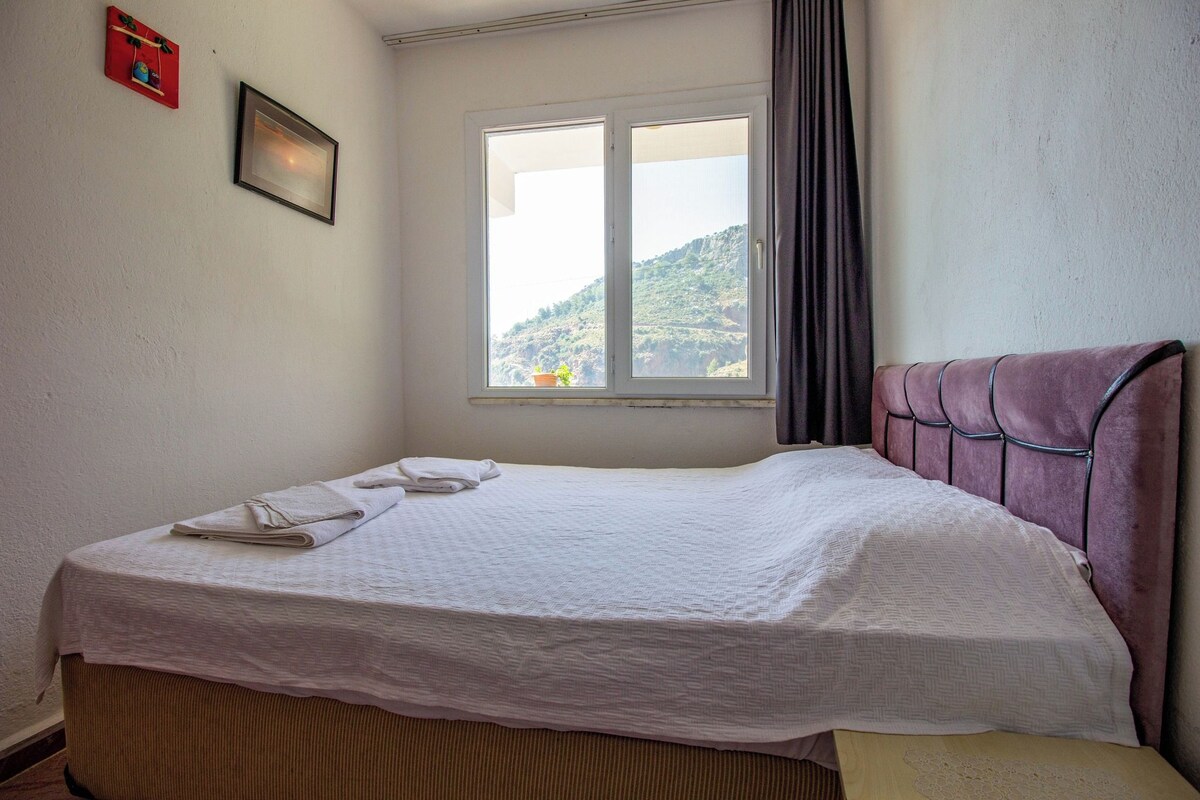 A neatly made bed with a white bedspread and two folded towels, a window with a view of a hillside, and a red wall decoration.
