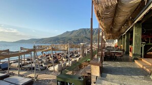Un restaurante frente a la playa con terraza, incluyendo sillones y mesas, con vistas al mar y a las montañas.
