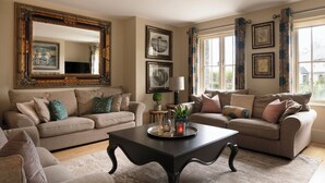 A living room with two beige sofas, a dark coffee table, and a large mirror. There are framed pictures on the walls and a window with curtains.
