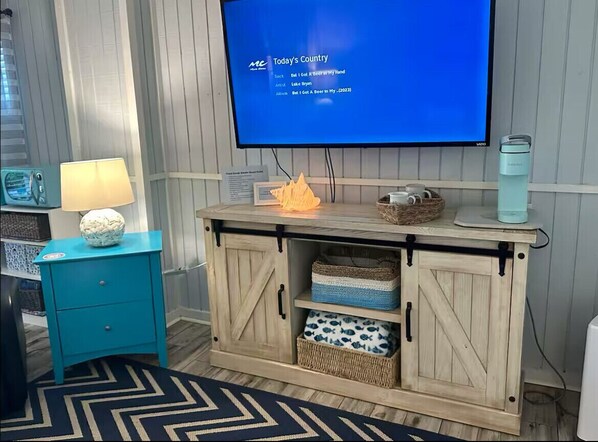 A room with a TV displaying a music album, a teal nightstand with a lamp, and a wooden cabinet with baskets.