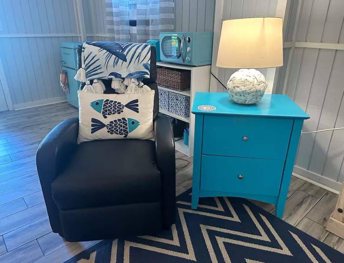 A room with a black armchair, a teal nightstand with a lamp, and a patterned rug.