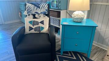 A room with a black armchair, a teal nightstand with a lamp, and a patterned rug.
