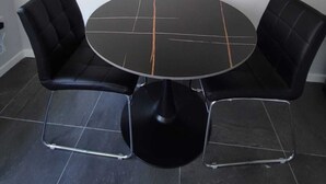 Ein runder Esstisch mit zwei schwarzen Polstersesseln und einer Bank, vor einer schlichten Wand mit einem gerahmten Gemälde darüber.