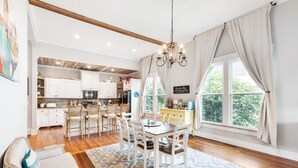 Une cuisine spacieuse avec des meubles blancs, un coin repas doté d’une table et de chaises en bois, et une grande fenêtre agrémentée de rideaux.