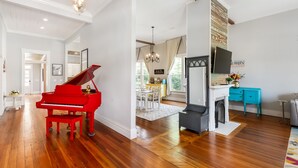A spacious living room with hardwood floors, a red grand piano, a fireplace, and a dining area with a chandelier. - Spacious 4-bedroom w/ full en suite baths -  1/2 mile from Covington Square! (Covington)