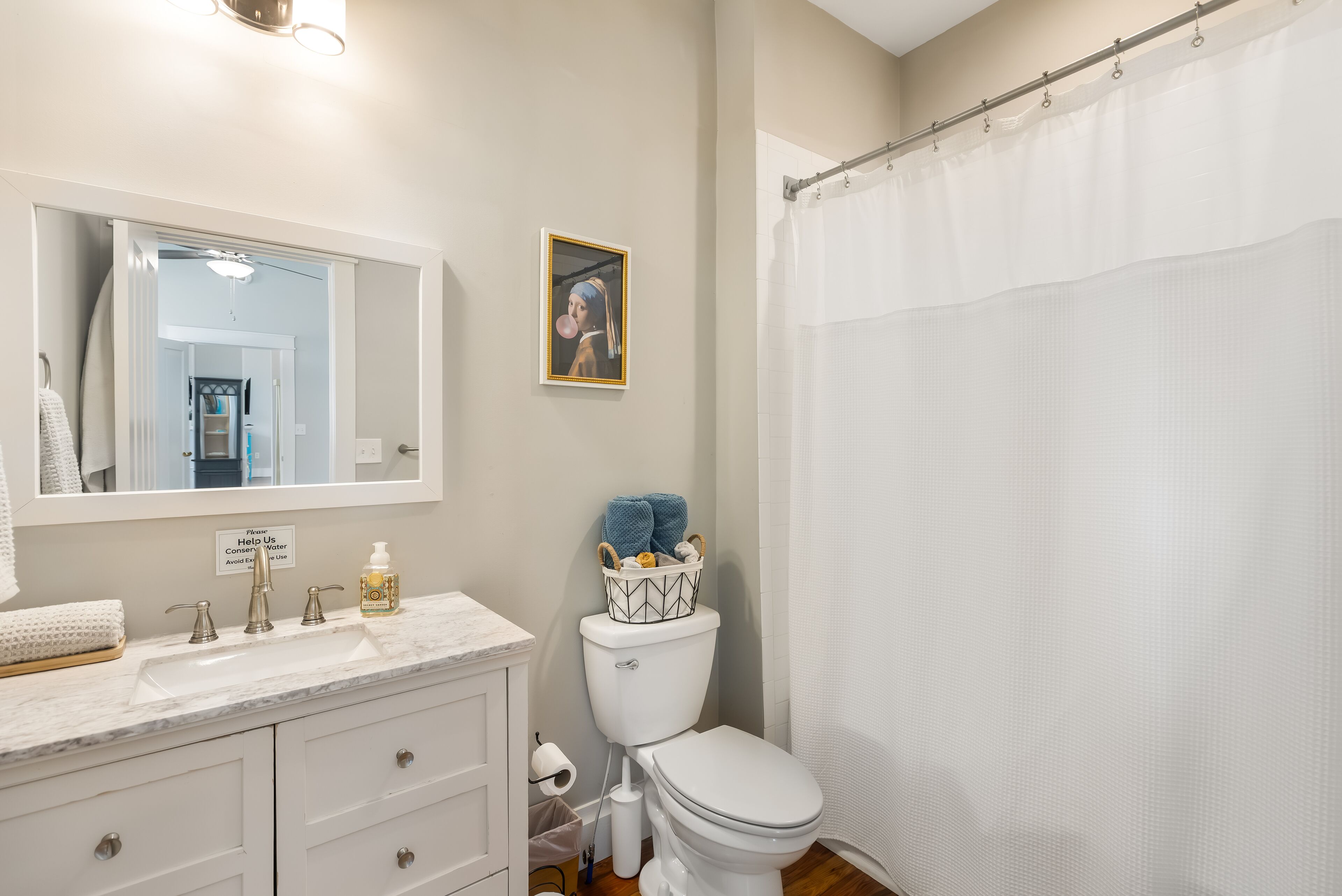 Un baño con lavamanos, espejo y retrete blancos. Tiene una cortina de ducha y un letrero sobre el inodoro.