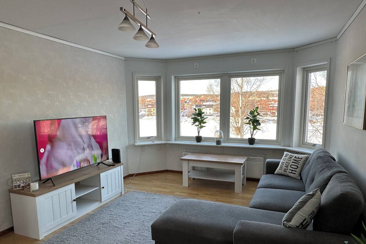 Uma sala de estar moderna com uma televisão de tela plana grande, um sofá cinza, uma mesa de centro de madeira e uma janela ampla com vista para edifícios.