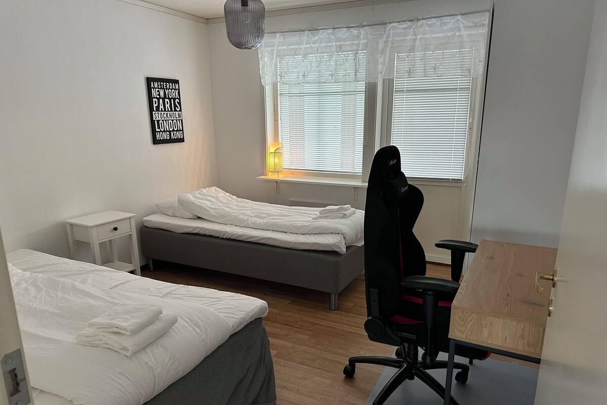 Quarto com duas camas, uma escrivaninha, uma cadeira, janela com persianas e um letreiro na parede listando cidades.