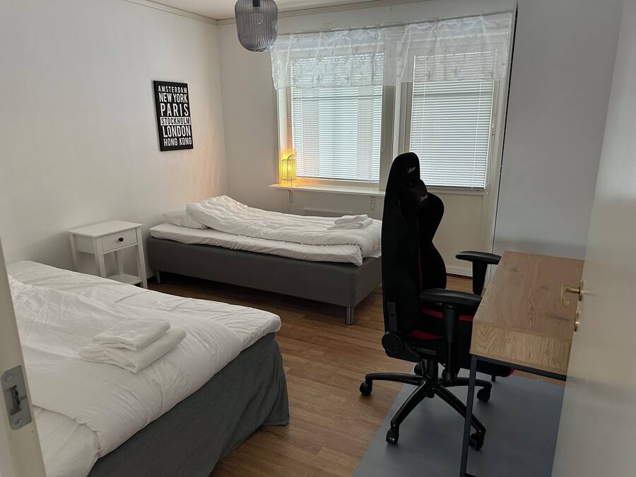 Ett sovrum med två sängar, ett skrivbord, en stol, ett fönster med persienner och en väggtavla med en lista över städer.