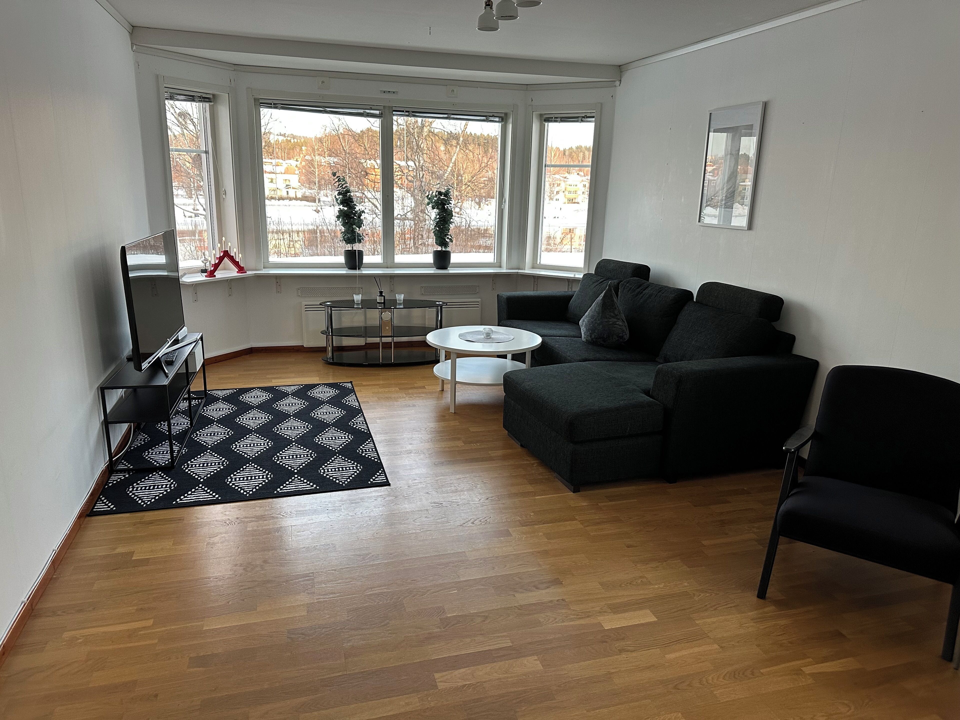 A modern living room with a black sofa, a white coffee table, and a TV on a stand. There are large windows with a view of trees and buildings outside.