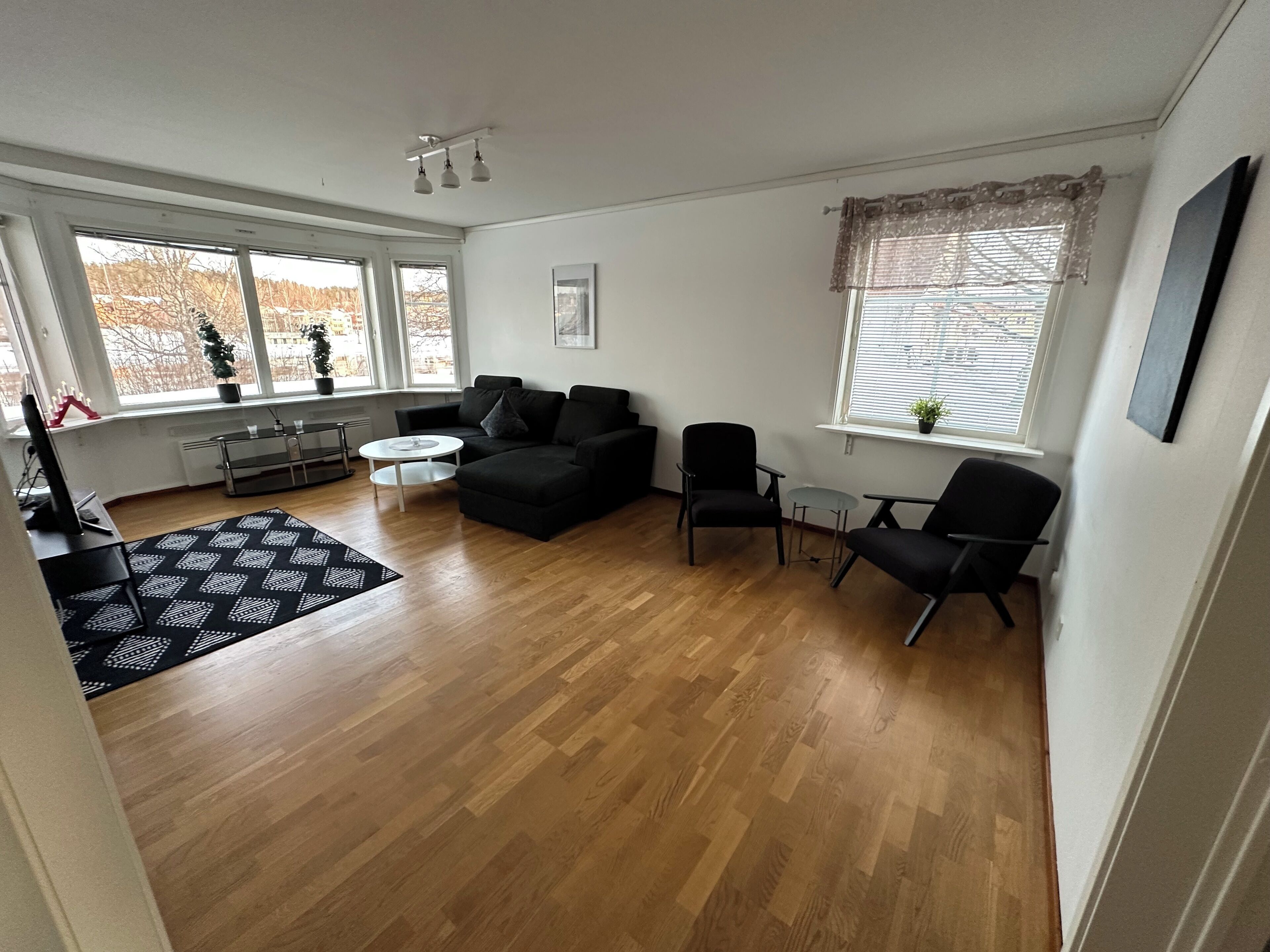 A modern living room with a black sofa, a coffee table, and two black armchairs. There are large windows with blinds, a radiator, and a small table with a potted plant.