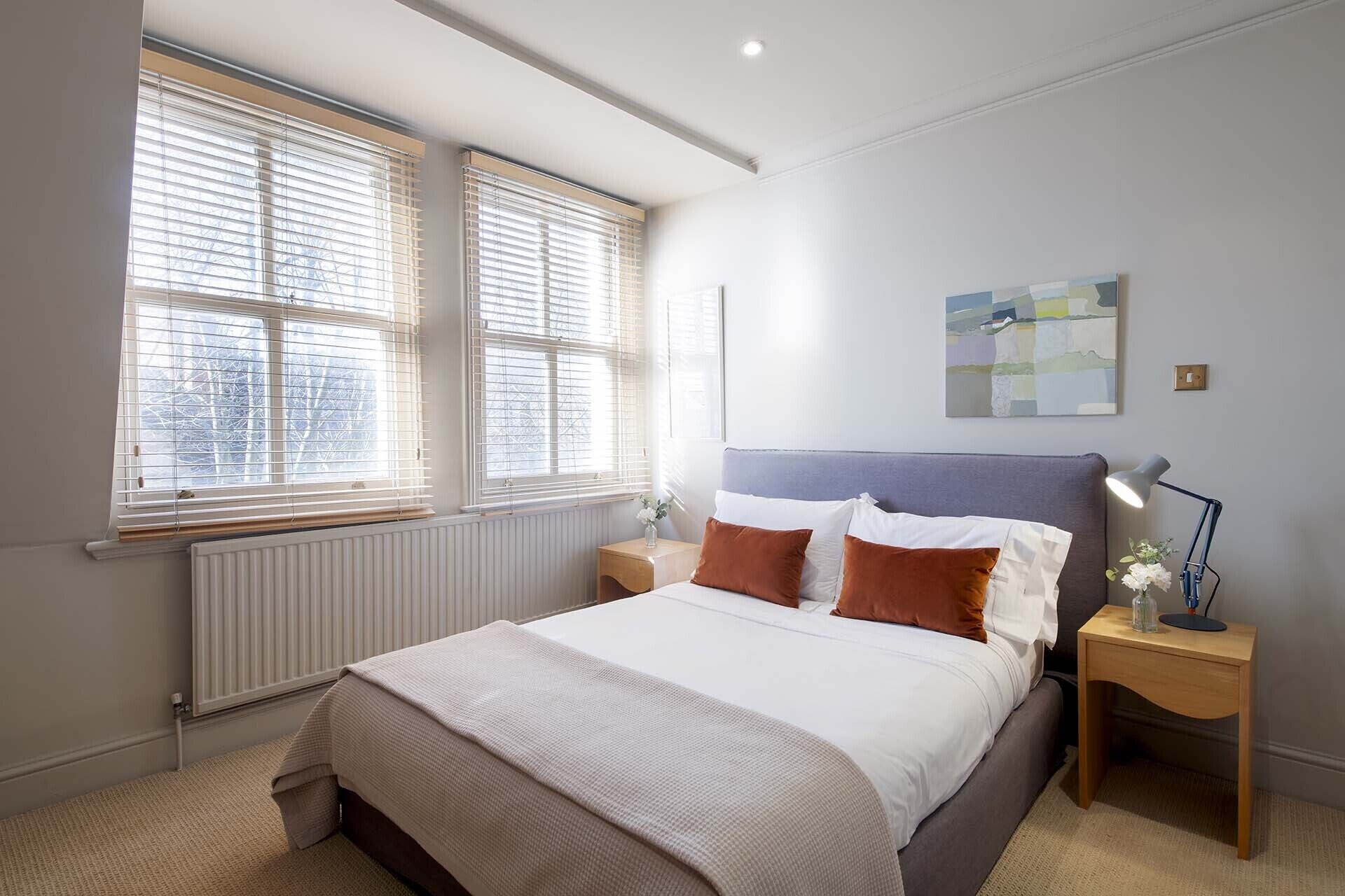 Ett sovrum med en säng, två sängbord, ett litet bord, en lampa och ett fönster med persienner.