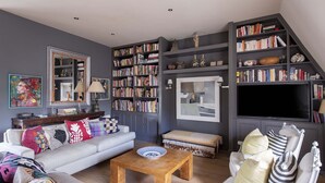 Ein Wohnzimmer mit grauem Sofa, einem Holztisch, einer Bücherwand voller Bücher, einem Flachbildfernseher und einem dekorativen Spiegel.