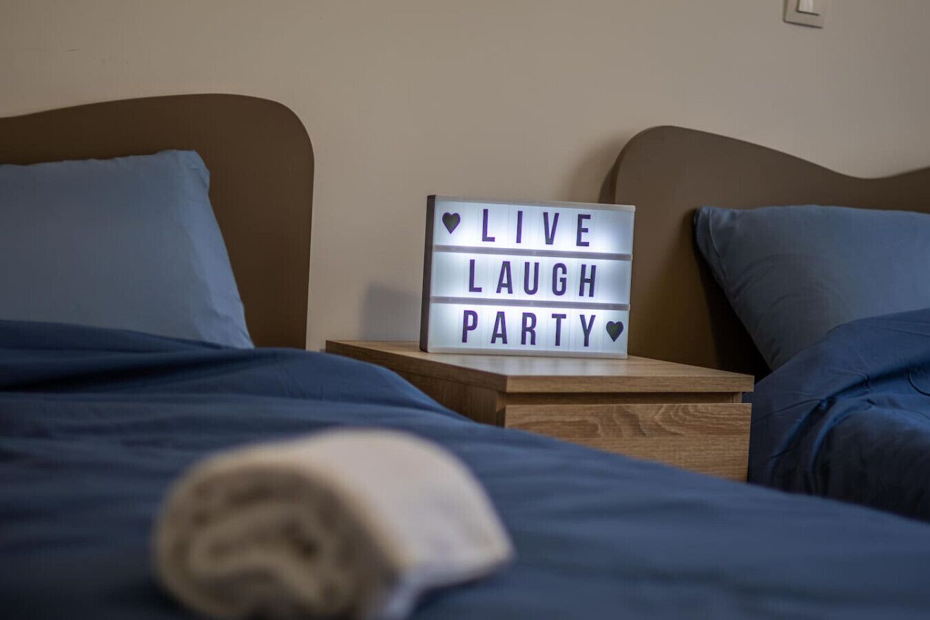 Ein ordentlich gemachtes Bett mit blauer Bettwäsche und ein lightbox-Schild mit der Aufschrift "LIVE LAUGH PARTY" auf einem Nachttisch aus Holz.
