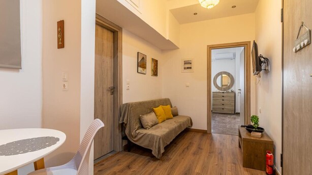 Una habitación acogedora con piso de madera, un sofá con cojines, una mesita y una silla. Se ve una puerta que da a otra habitación y un espejo en la pared.