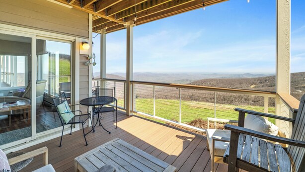 Une terrasse en bois avec des meubles d’extérieur, une porte vitrée donnant sur l’intérieur et une vue sur un paysage vallonné.