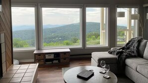 Un salon moderne doté d’une grande fenêtre donnant sur des collines verdoyantes. On y trouve un canapé, une table basse avec un carnet et des lunettes, ainsi qu’une petite table d’appoint.