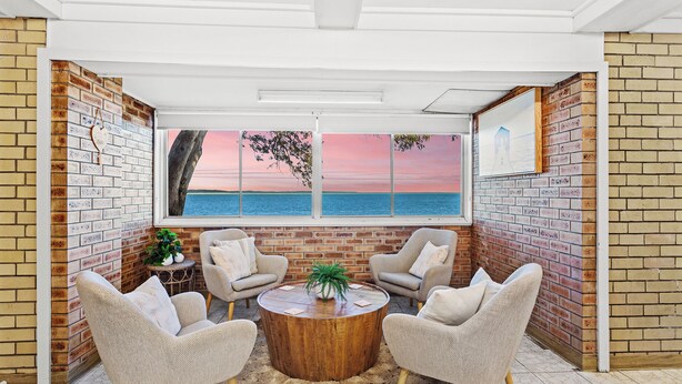 A room with a brick wall, a large window with a sunset view, two armchairs, a round wooden coffee table, and a small plant. - Absolute waterfront property on rock wall (Amity Point)