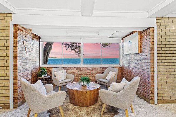 A room with a brick wall, a large window with a sunset view, two armchairs, a round wooden coffee table, and a small plant.