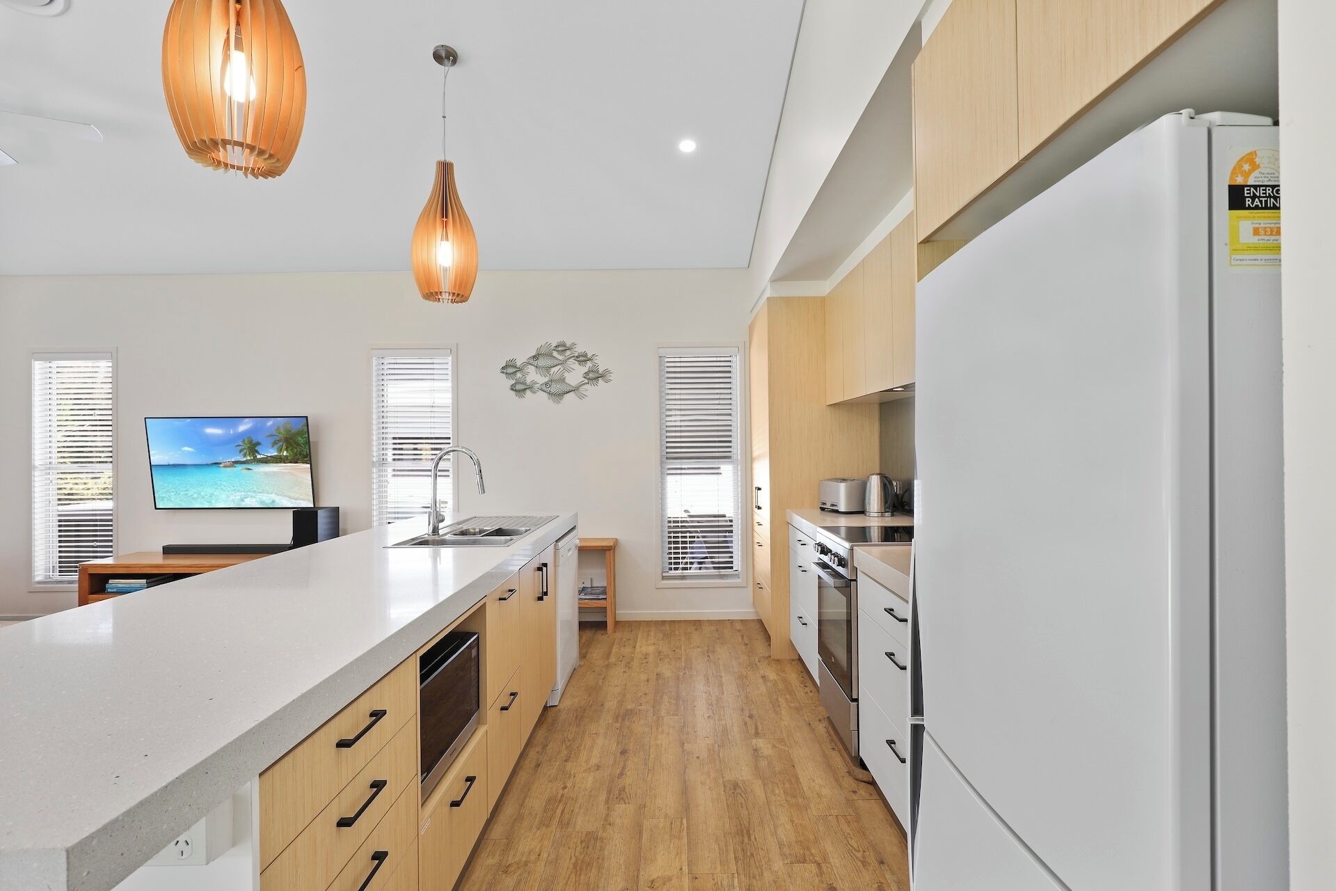 A modern kitchen with a white refrigerator, wooden cabinets, and a large island with a sink. There are two pendant lights and a TV mounted on the wall.