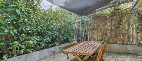 A wooden picnic table with benches under a shaded area, surrounded by lush greenery and bamboo.