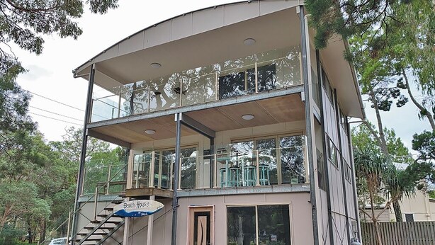Un edificio de dos pisos con un balcón en el piso superior, una escalera que conduce a la entrada y un letrero que indica que es una casa de playa.